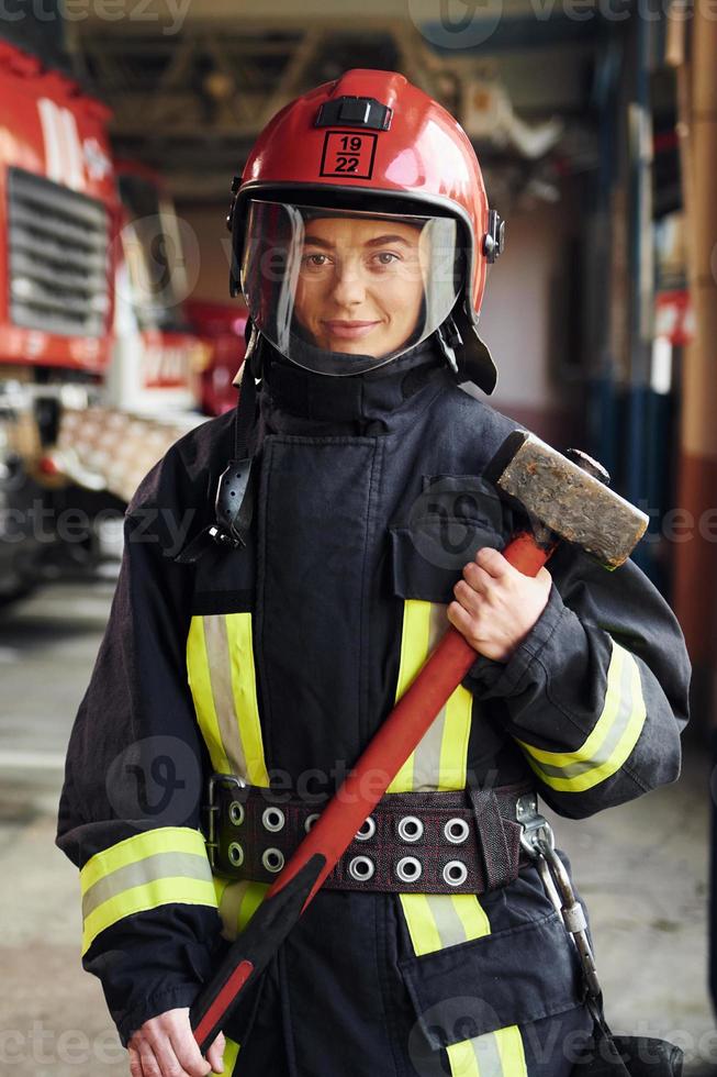 con martillo en las manos. bombero femenino en uniforme protector de pie cerca de camión foto