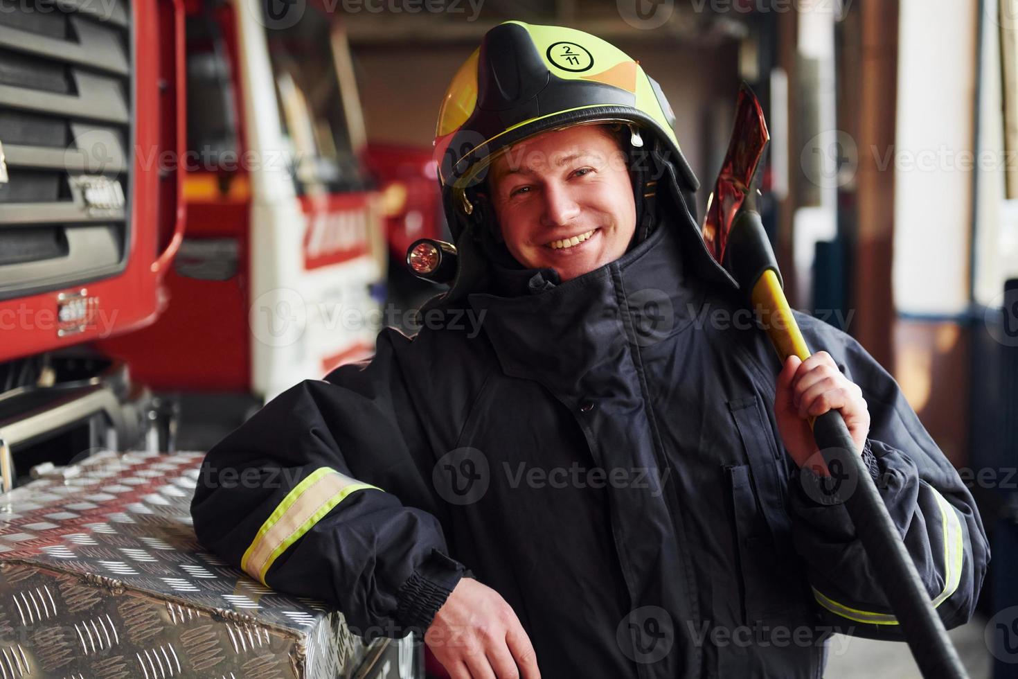 Holds axe in hands. Male firefighter in protective uniform standing near truck photo