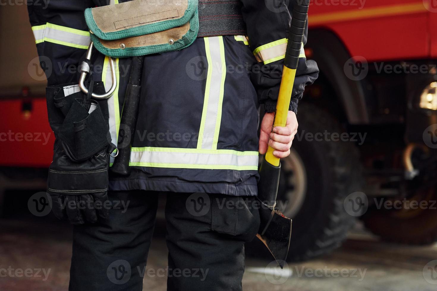 Holds axe in hands. Male firefighter in protective uniform standing near truck photo