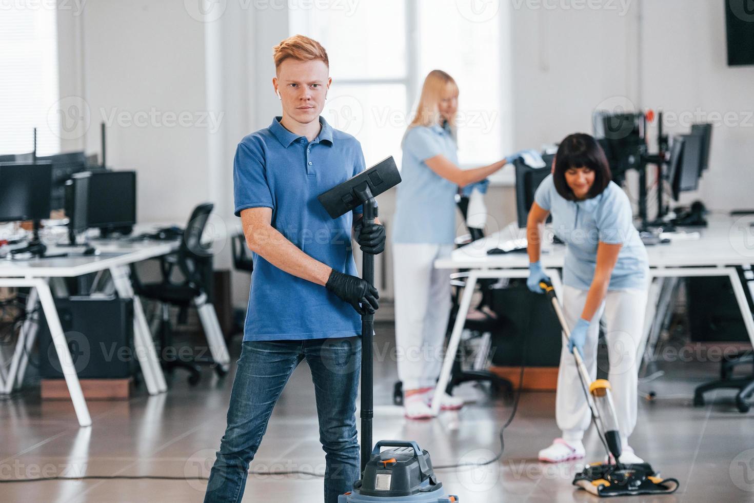 Guy with vacuum cleaner. Group of workers clean modern office together at daytime photo