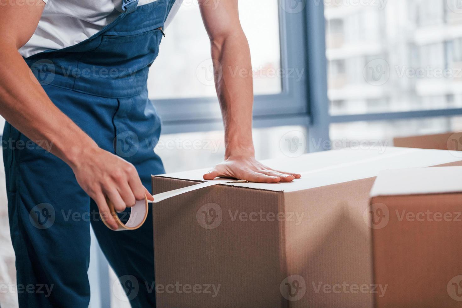 Packaging the box. Young male mover in blue uniform works indoors in the room photo