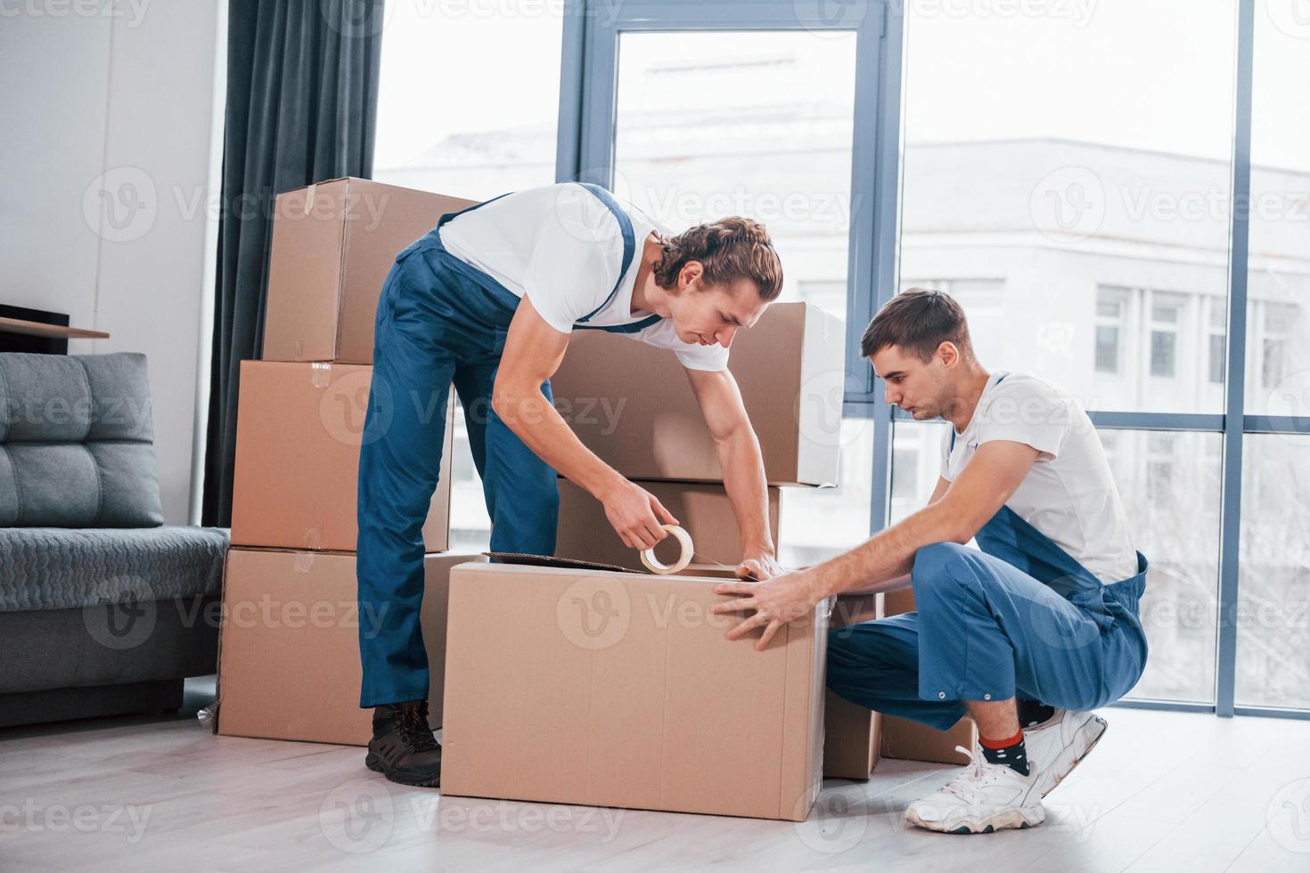 Packaging the box. Two young movers in blue uniform working indoors in the room photo