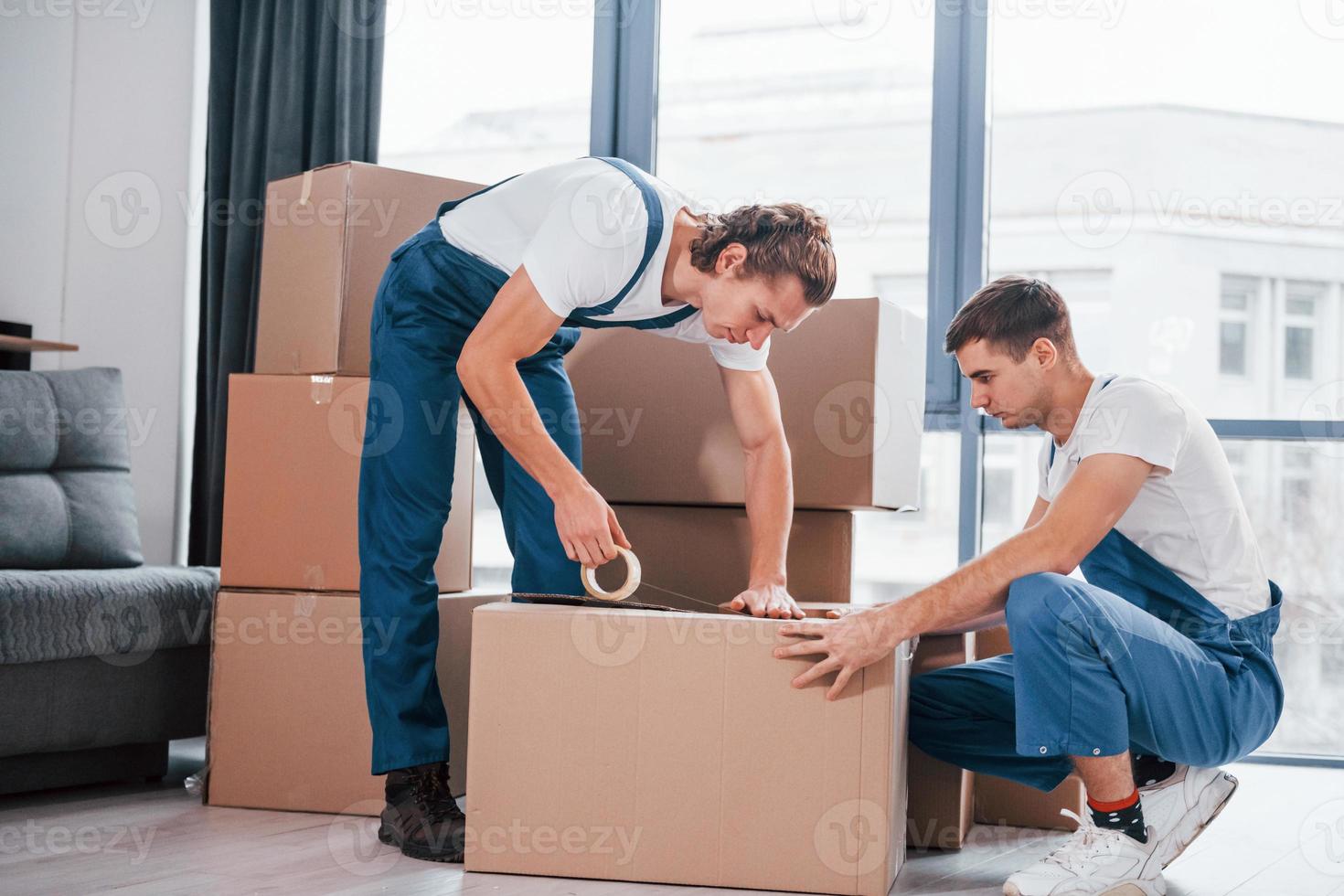 Packaging the box. Two young movers in blue uniform working indoors in the room photo