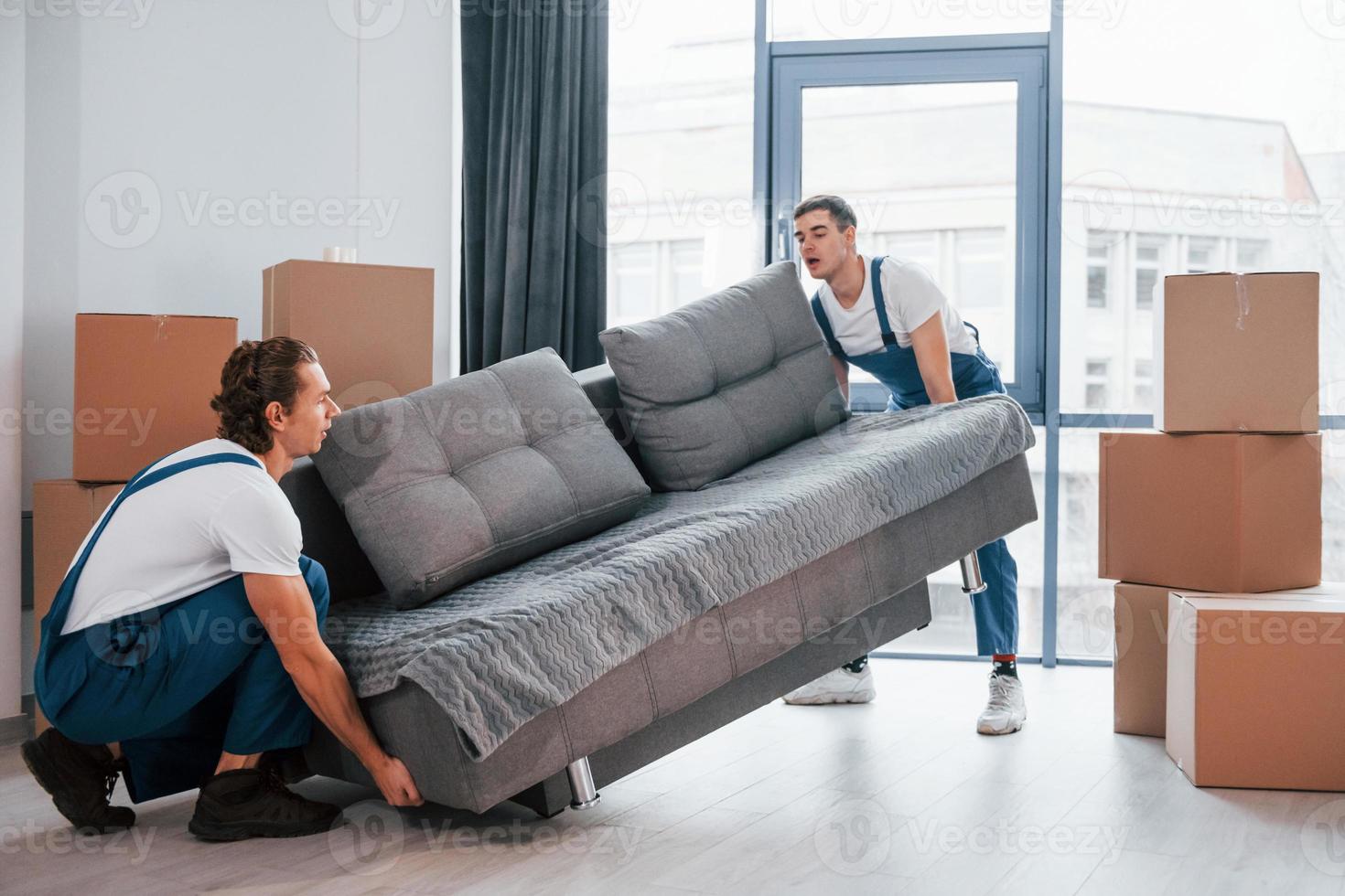 Carrying heavy sofa. Two young movers in blue uniform working indoors