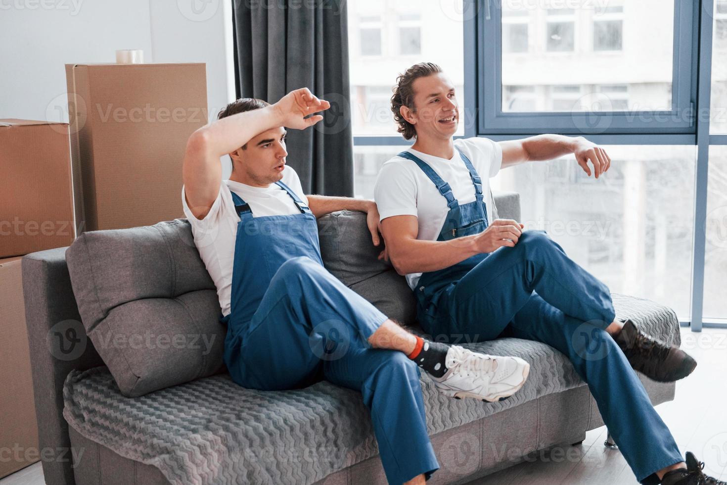 Sitting on the sofa. Tired and taking break. Two young movers in blue uniform working indoors in the room photo