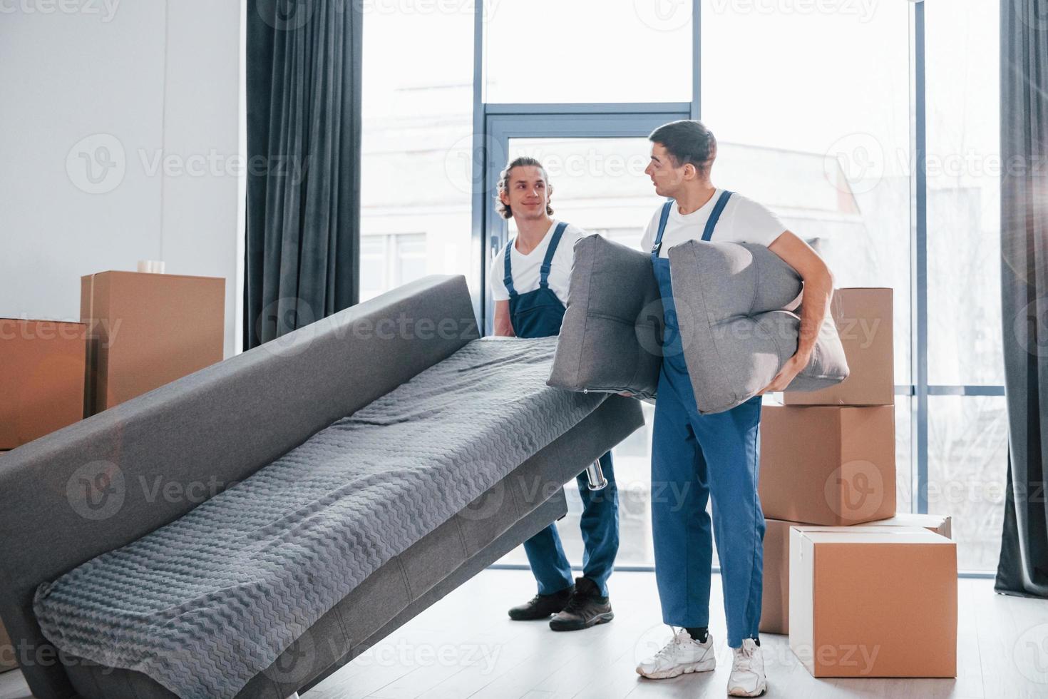 Carrying heavy sofa. Two young movers in blue uniform working indoors in the room photo