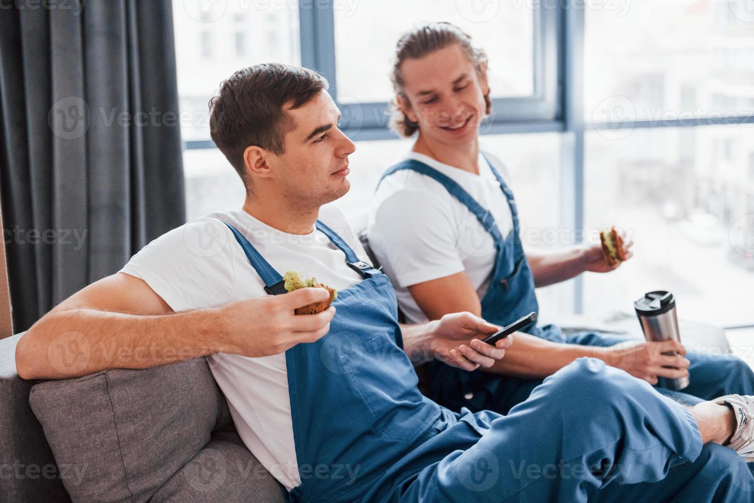 Sitting on the sofa. Tired and taking break. Two young movers in blue uniform working indoors in the room photo