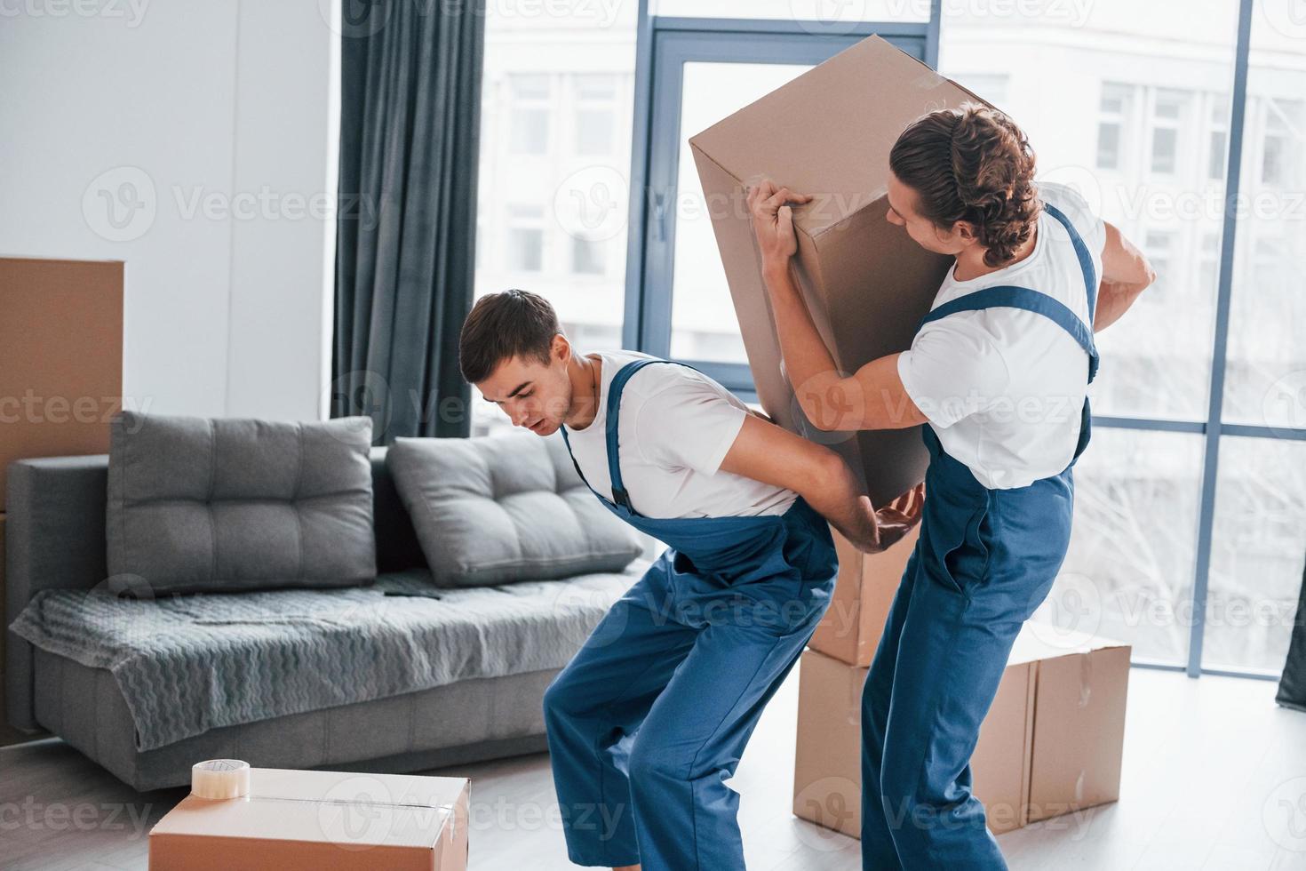 Heavy boxes. Two young movers in blue uniform working indoors in the room photo