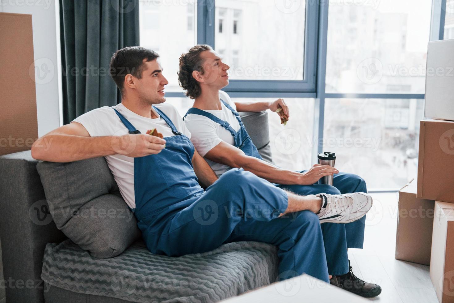 Sitting on the sofa. Tired and taking break. Two young movers in blue uniform working indoors in the room photo