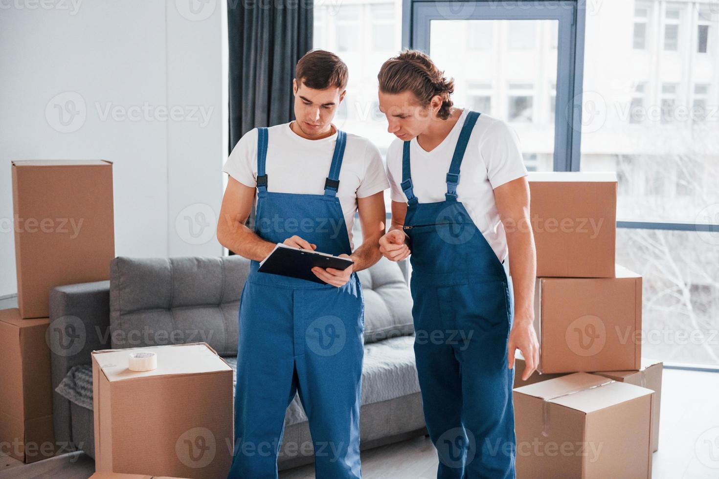 Holds notepad with document. Two young movers in blue uniform working indoors in the room photo