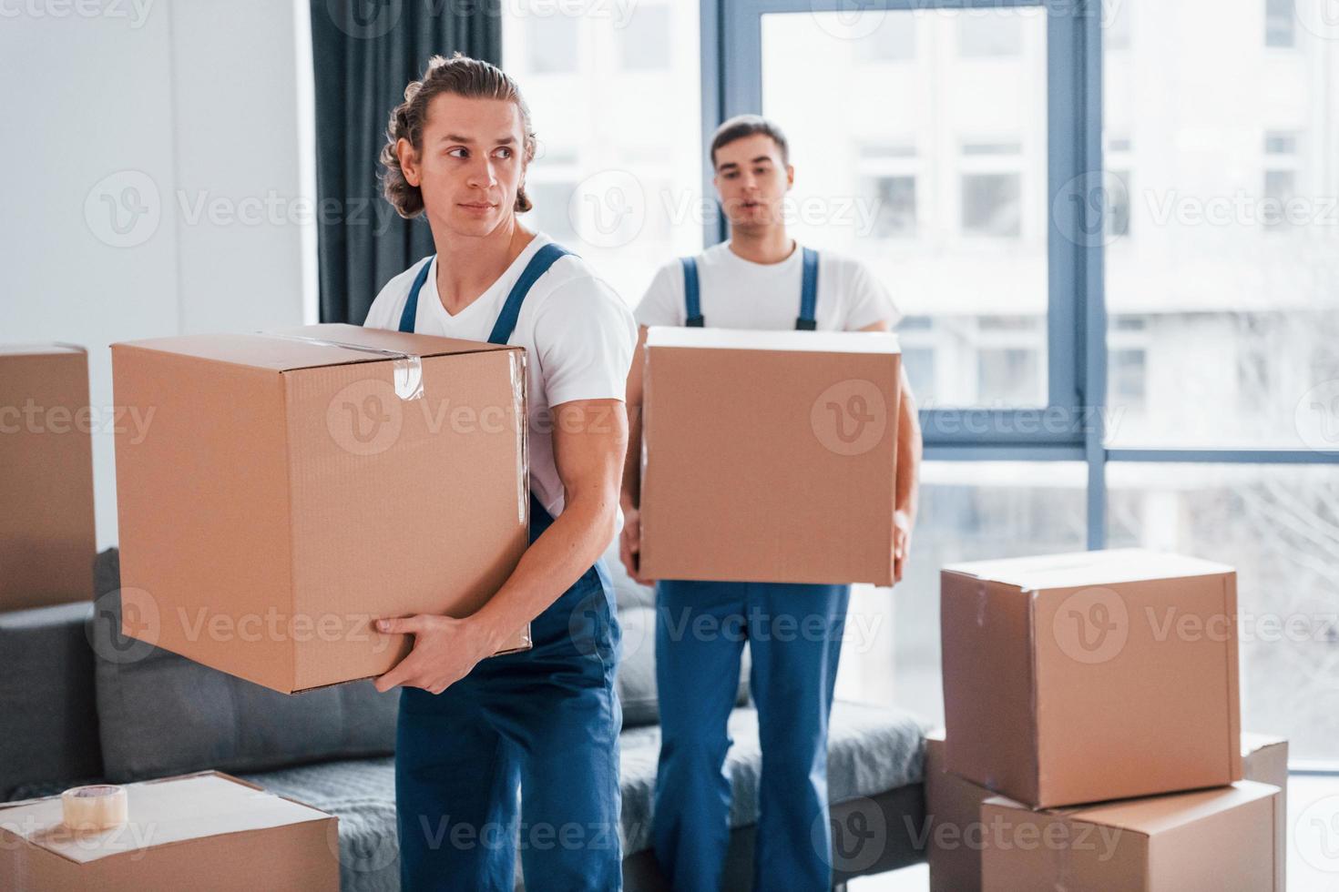 With boxes in hands. Two young movers in blue uniform working indoors in the room photo