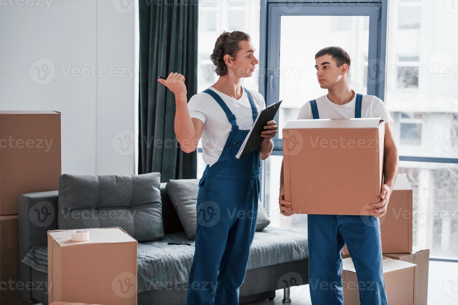 Holds notepad with document. Two young movers in blue uniform working indoors in the room photo
