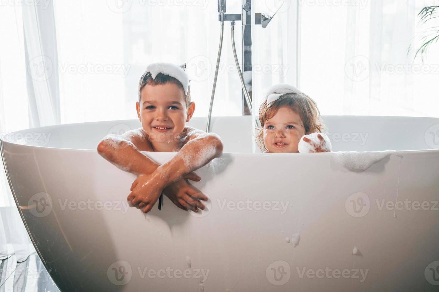 Two kids having fun and washing themselves in the bath at home. Posing for a camera photo