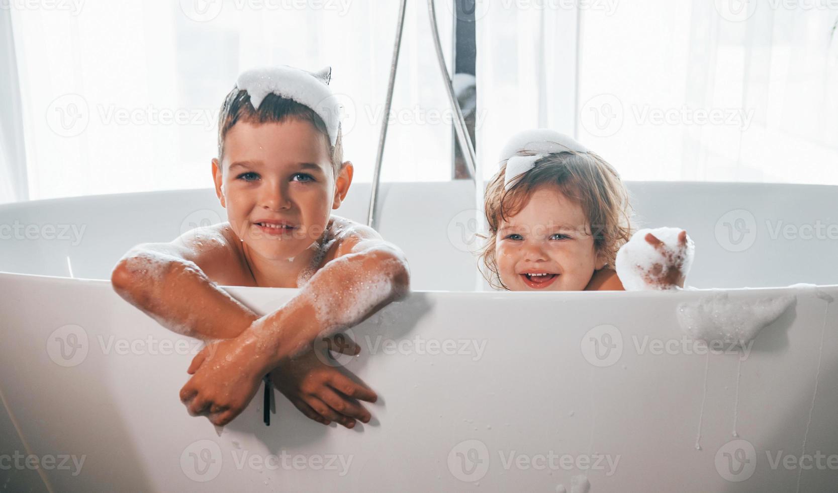 Two kids having fun and washing themselves in the bath at home. Posing for a camera photo