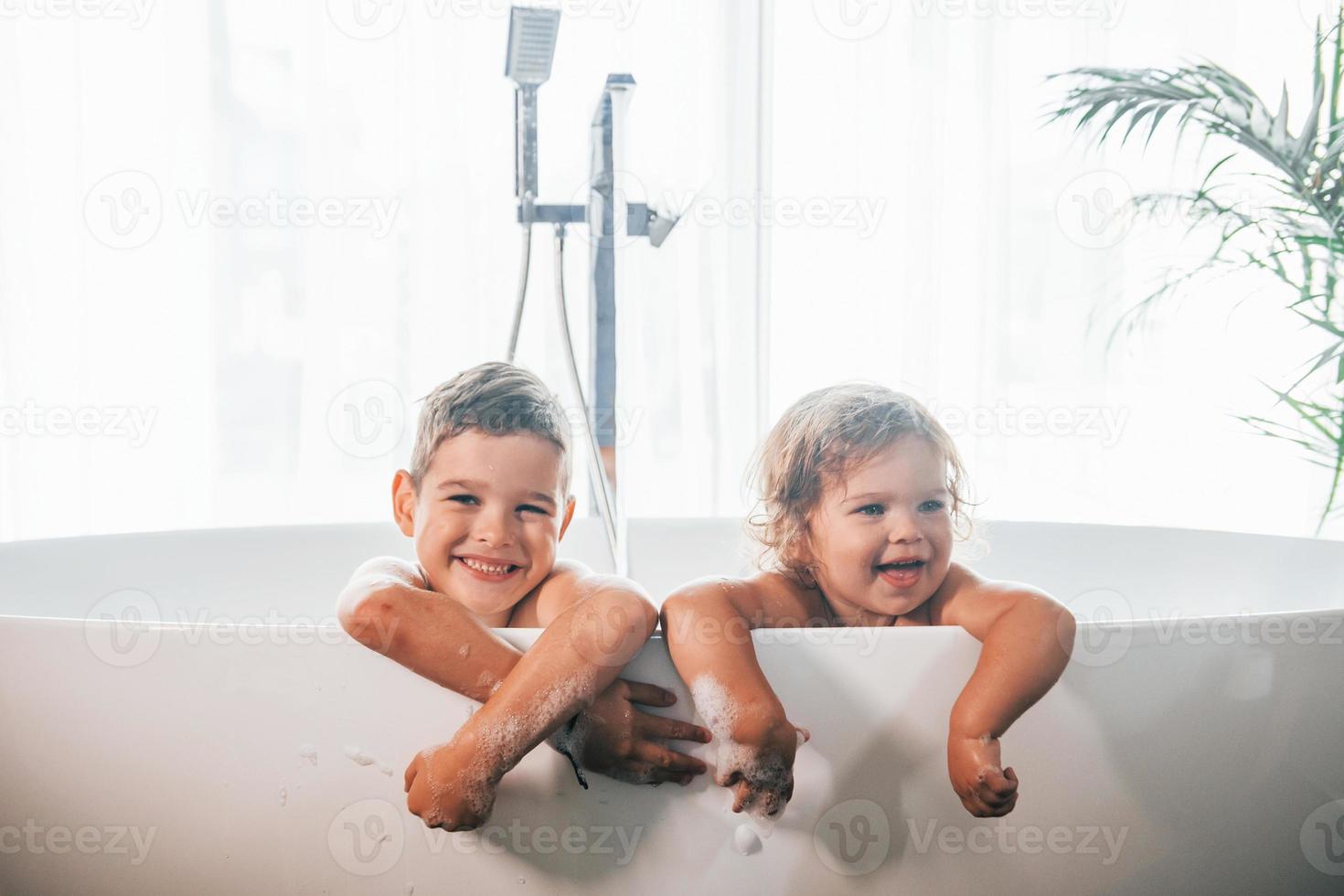 Two kids having fun and washing themselves in the bath at home. Posing for a camera photo