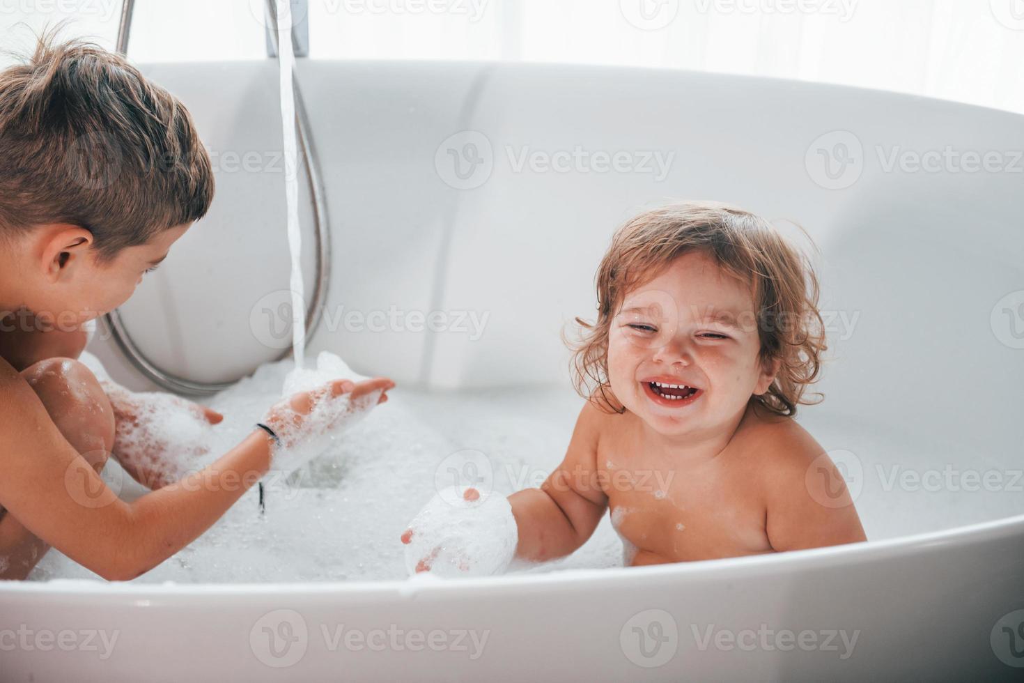 Two kids having fun and washing themselves in the bath at home. Posing for a camera photo