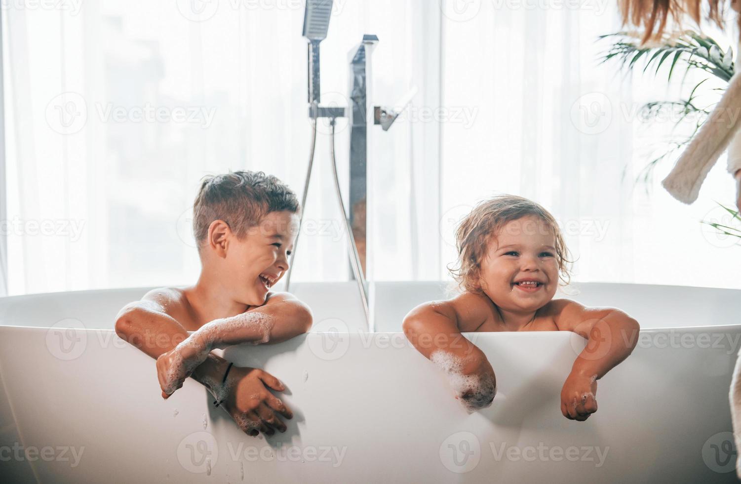 Two kids having fun and washing themselves in the bath at home. Posing for a camera photo