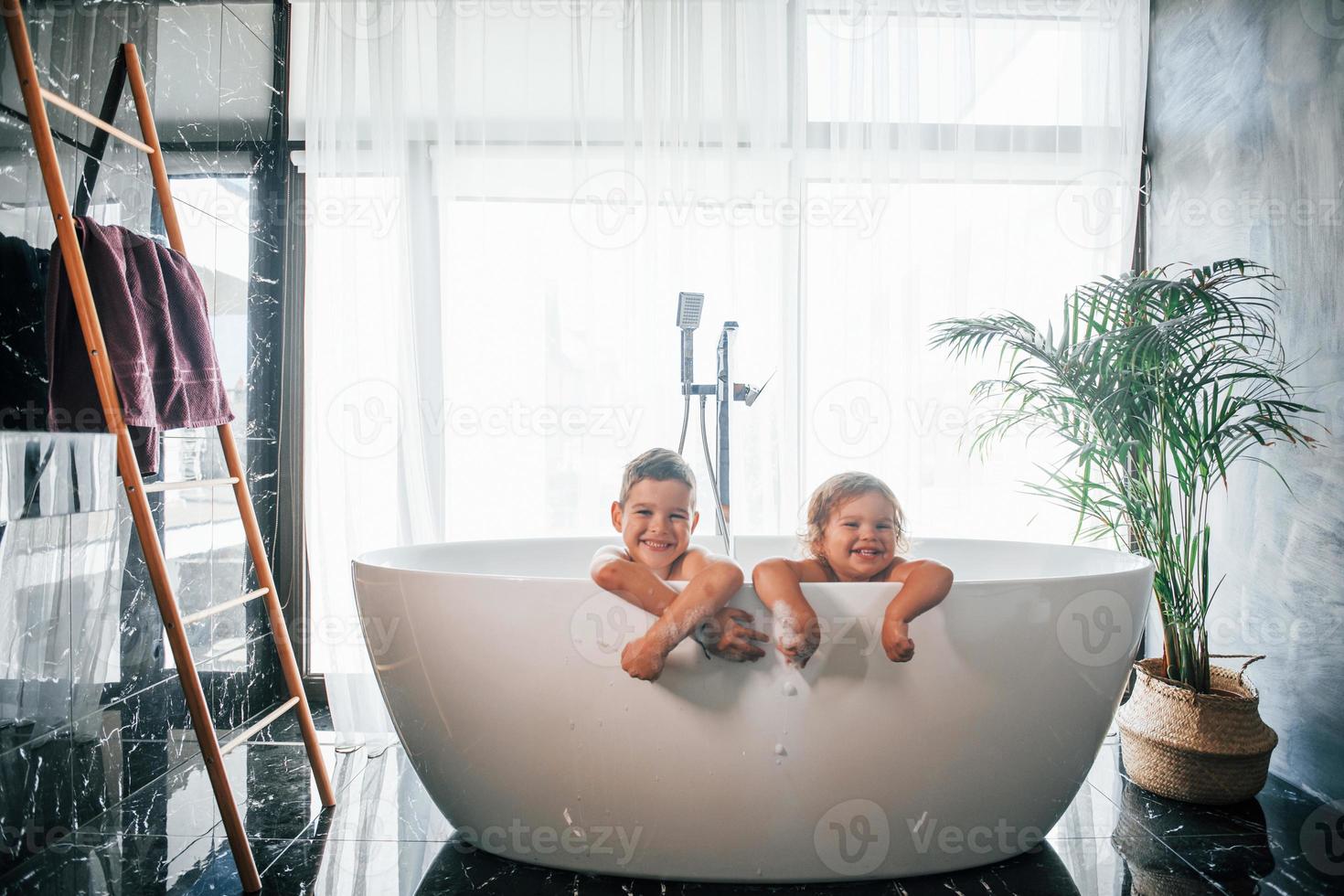 Two kids having fun and washing themselves in the bath at home. Posing for a camera photo