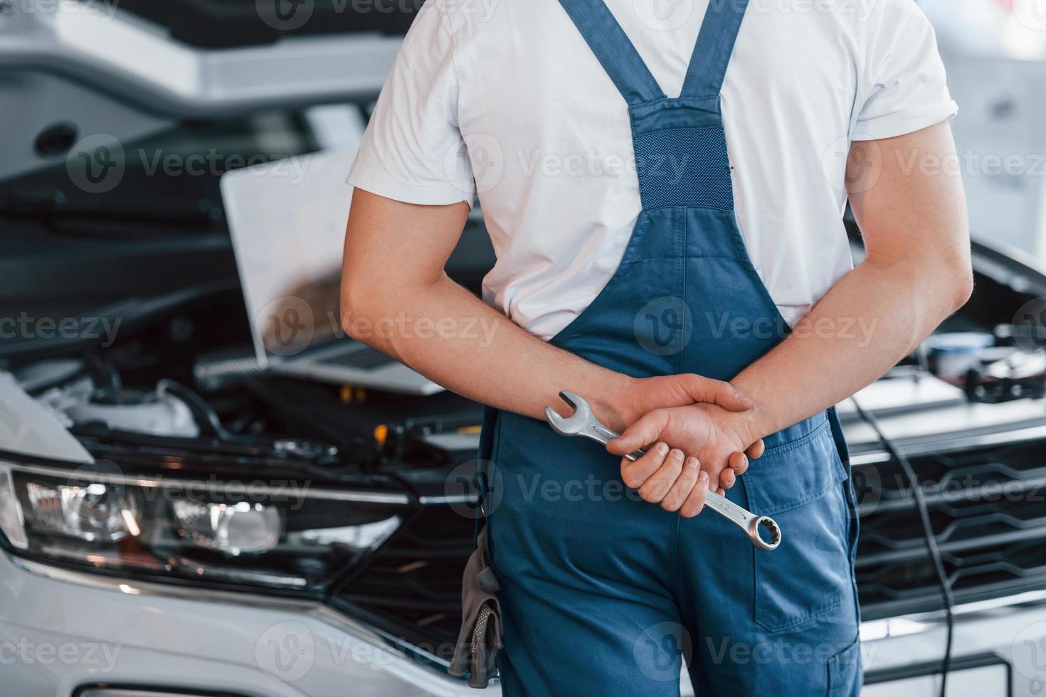 Uses laptop. Young man in white shirt and blue uniform repairs automobile photo