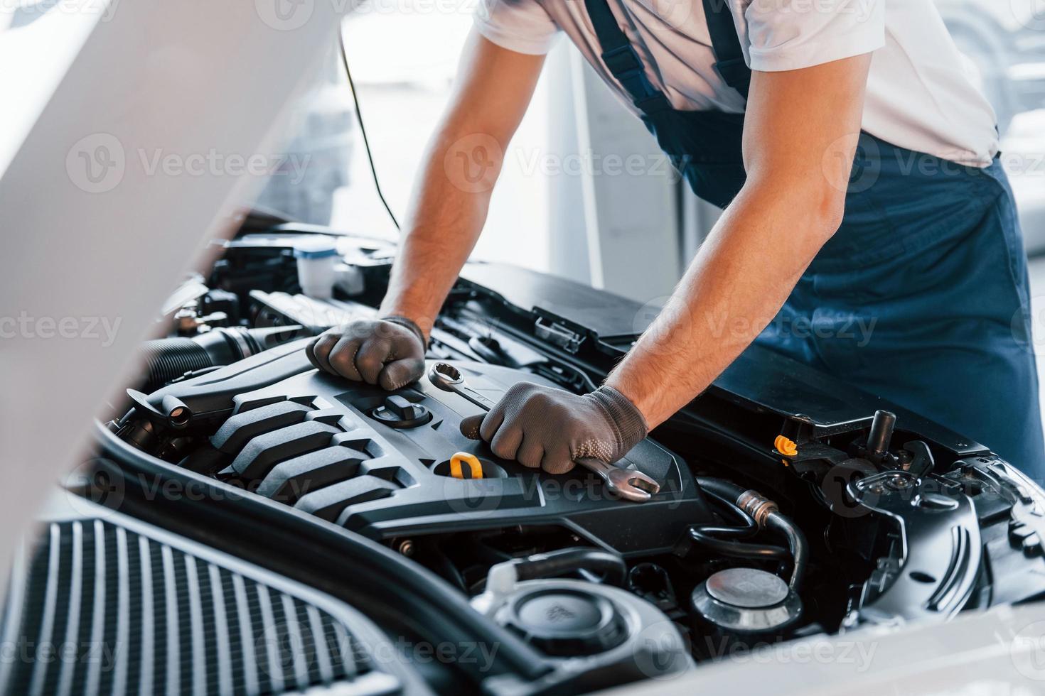 Quality service. Young man in white shirt and blue uniform repairs automobile photo