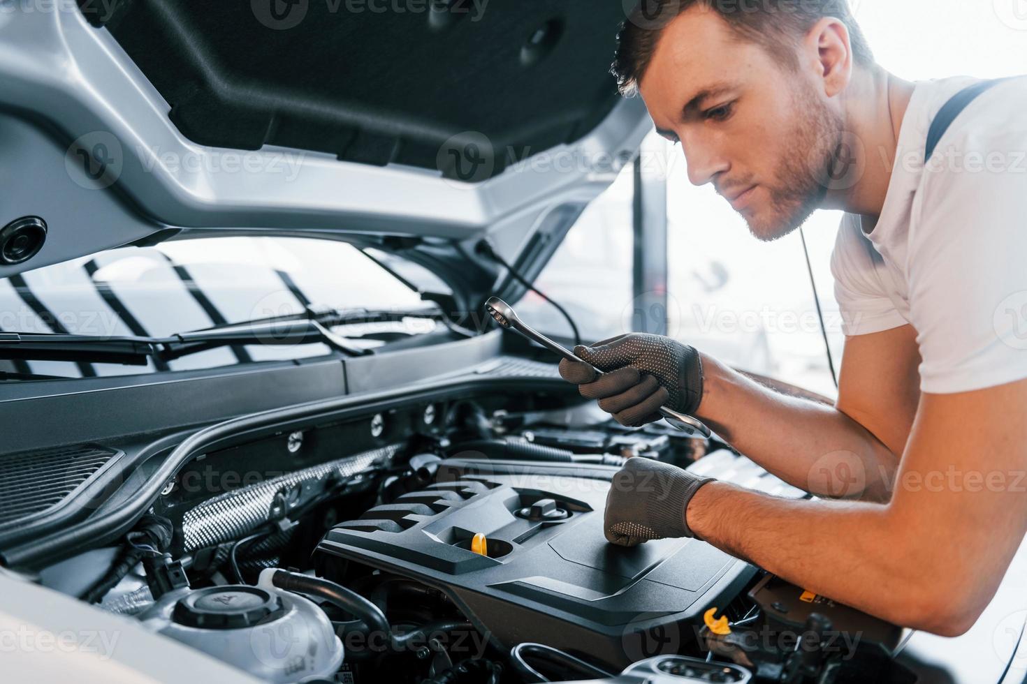 Quality service. Young man in white shirt and blue uniform repairs automobile photo
