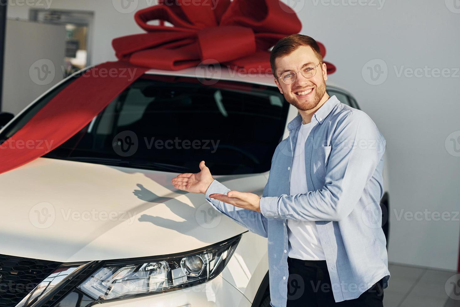 el automóvil está cubierto con cinta de regalo. hombre exitoso con anteojos parado cerca de un auto nuevo en el interior foto