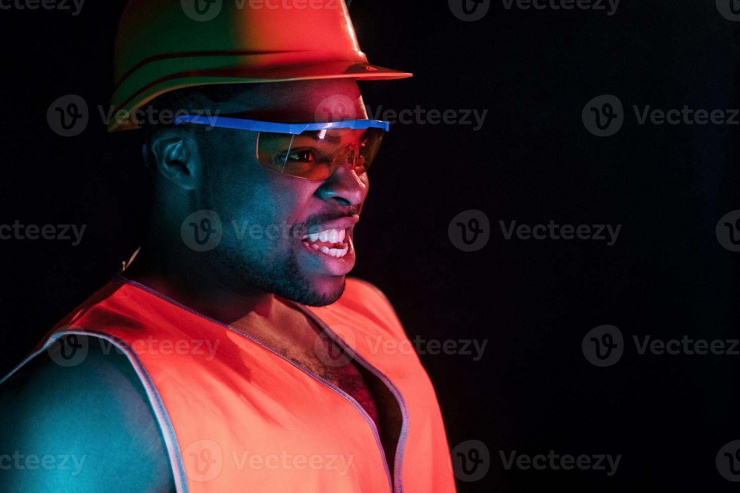 Construction worker in uniform and hard hat. Futuristic neon lighting. Young african american man in the studio photo