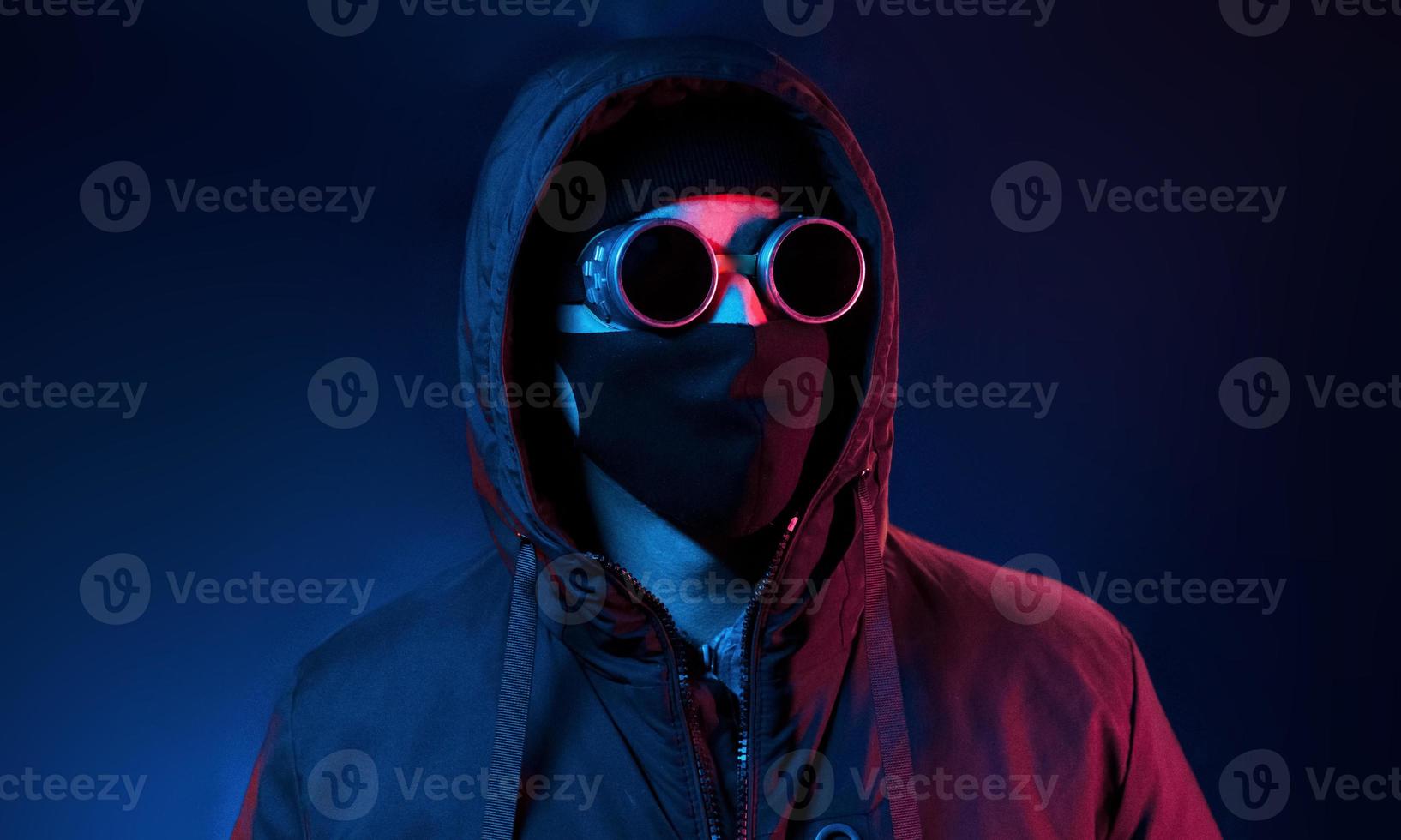 In mask, eyewear and hood. Neon lighting. Young european man is in the dark studio photo