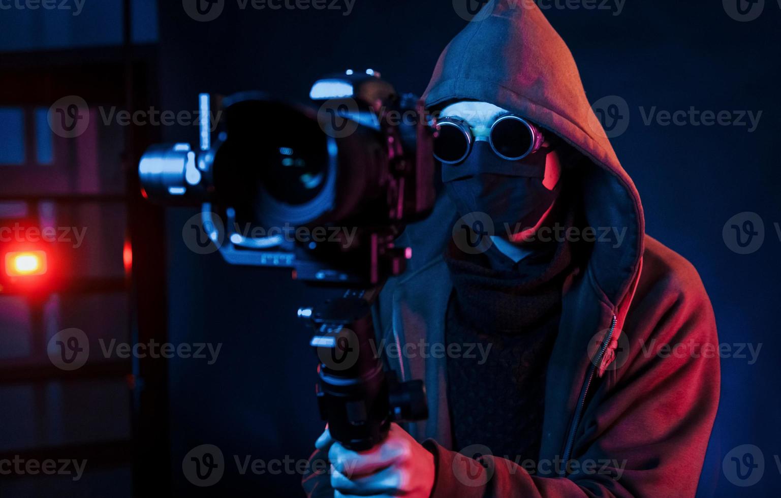 Guy in hood with camera. Neon lighting. Young european man is in the dark studio photo