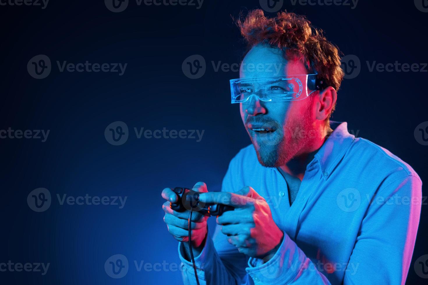 Plays game by using controller. Neon lighting. Young european man is in the dark studio photo