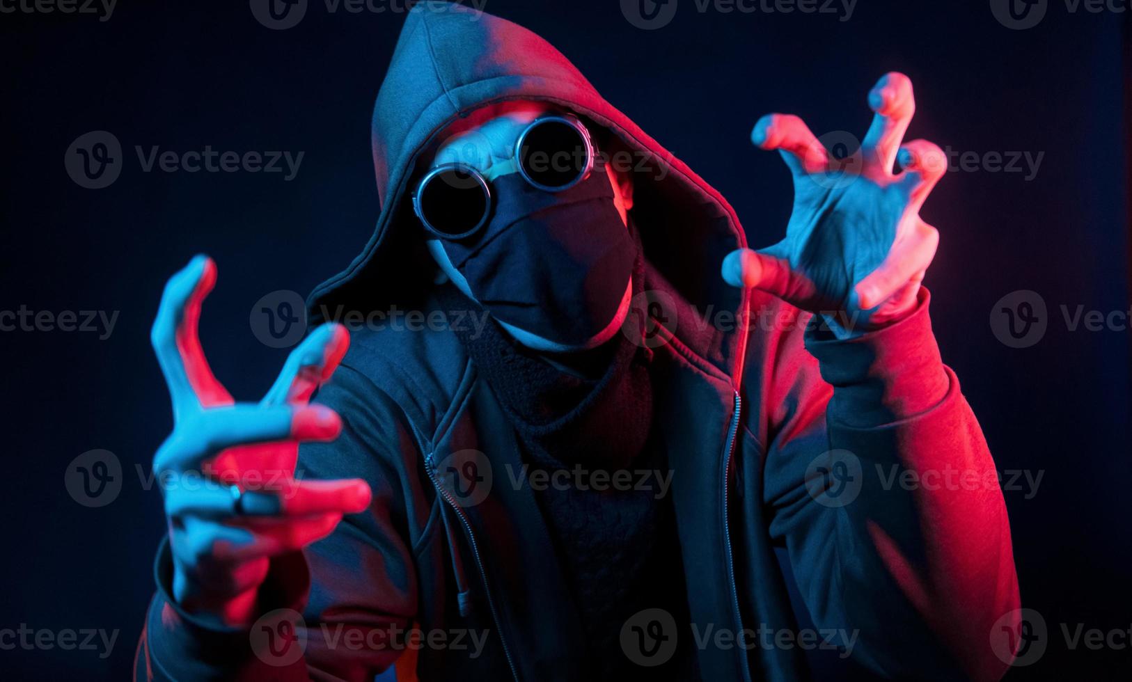 Scary guy in eyewear, mask and hood. Neon lighting. Young european man is in the dark studio photo