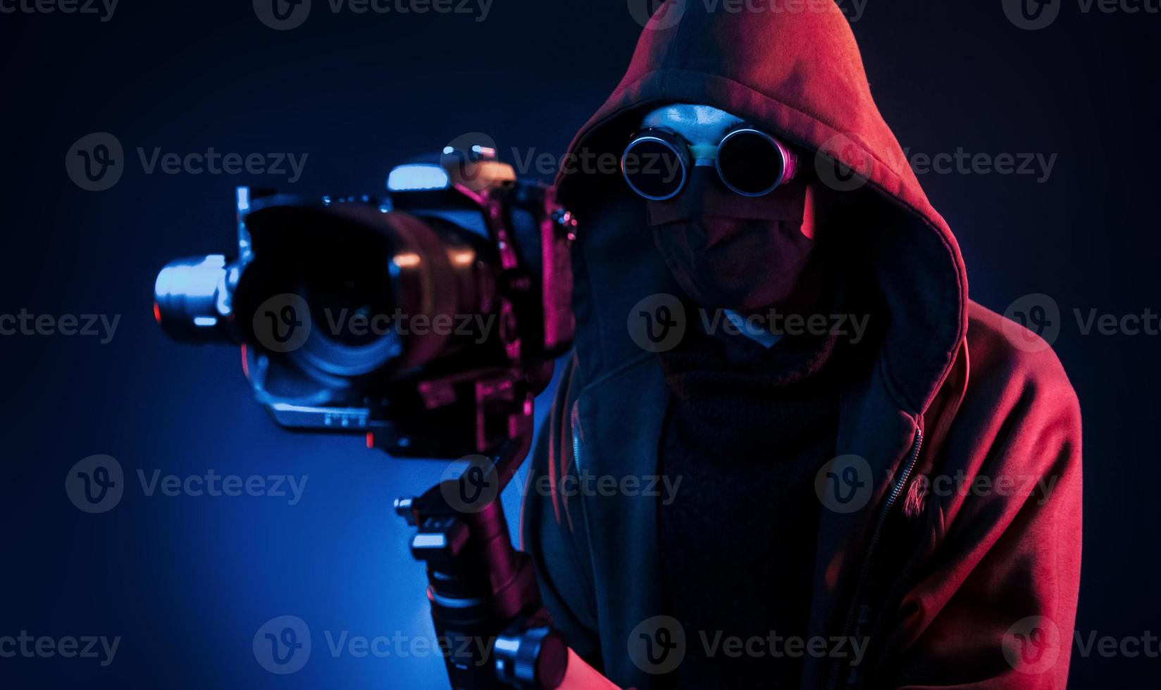 Guy in hood with camera. Neon lighting. Young european man is in the dark studio photo