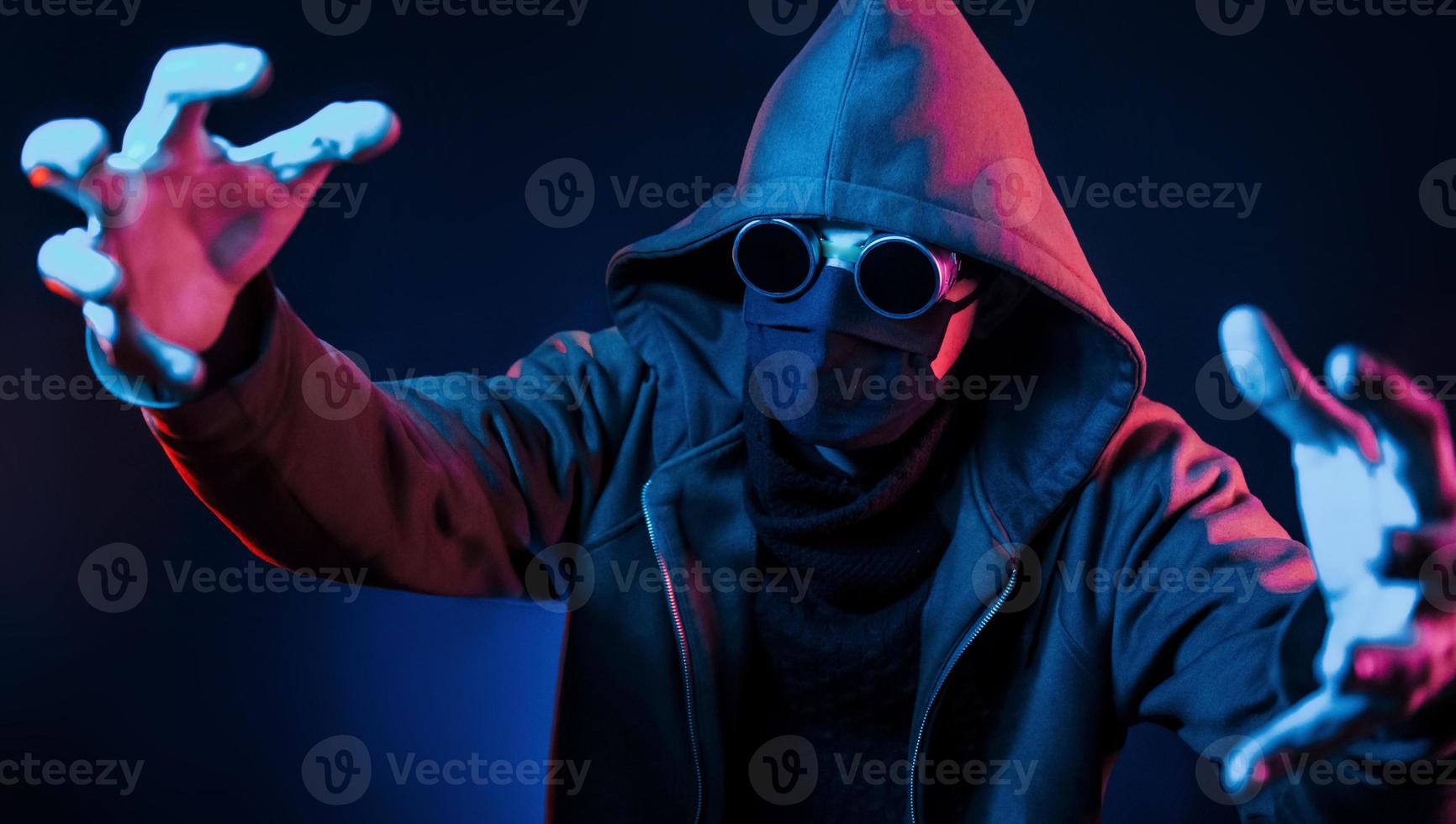 Scary guy in eyewear, mask and hood. Neon lighting. Young european man is in the dark studio photo