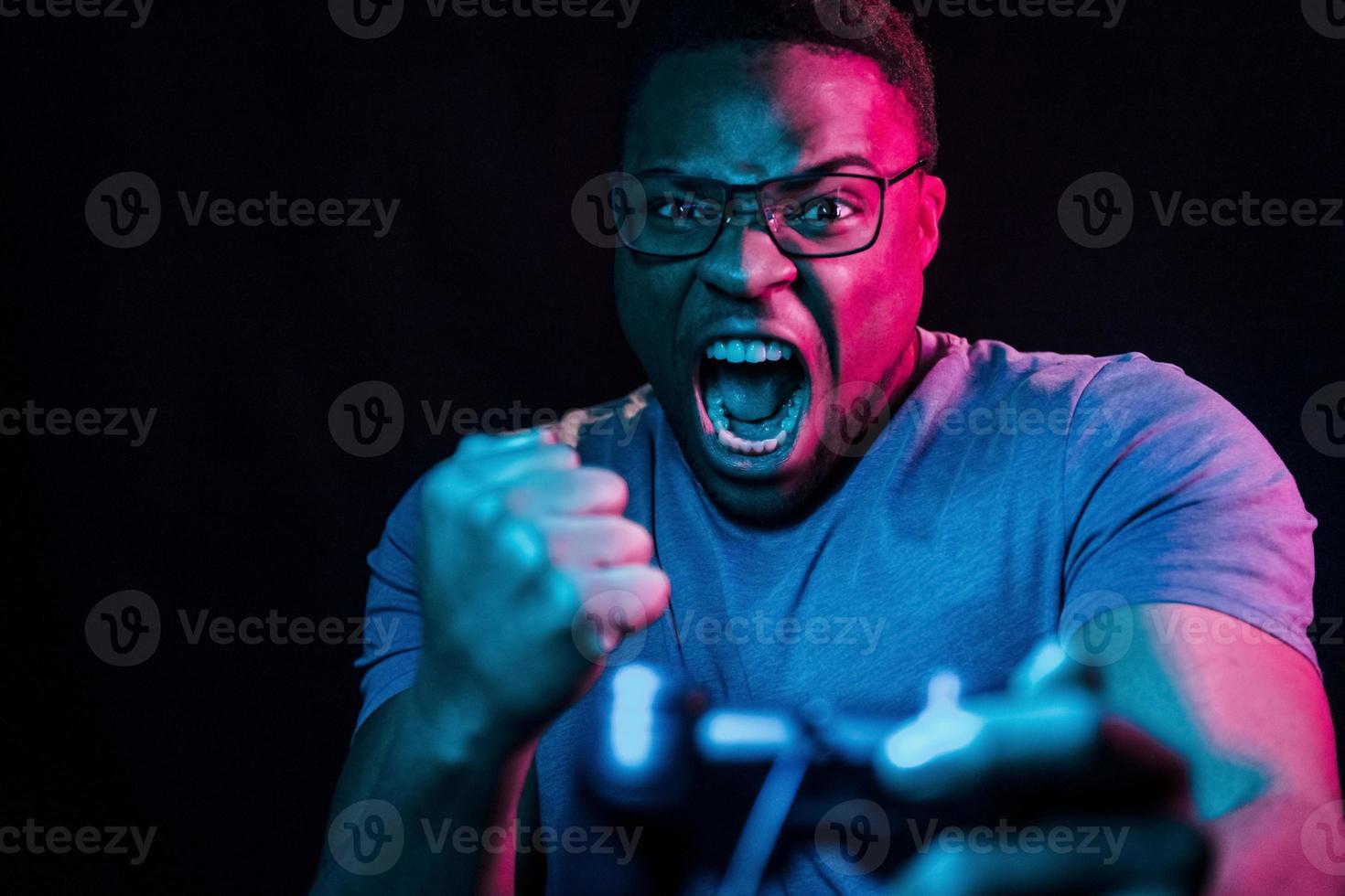 Plays game by using controller. Futuristic neon lighting. Young african american man in the studio photo