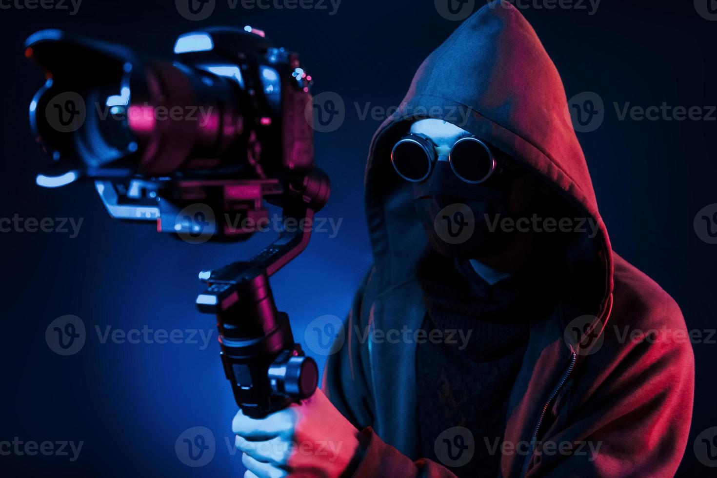 Guy in hood with camera. Neon lighting. Young european man is in the dark studio photo