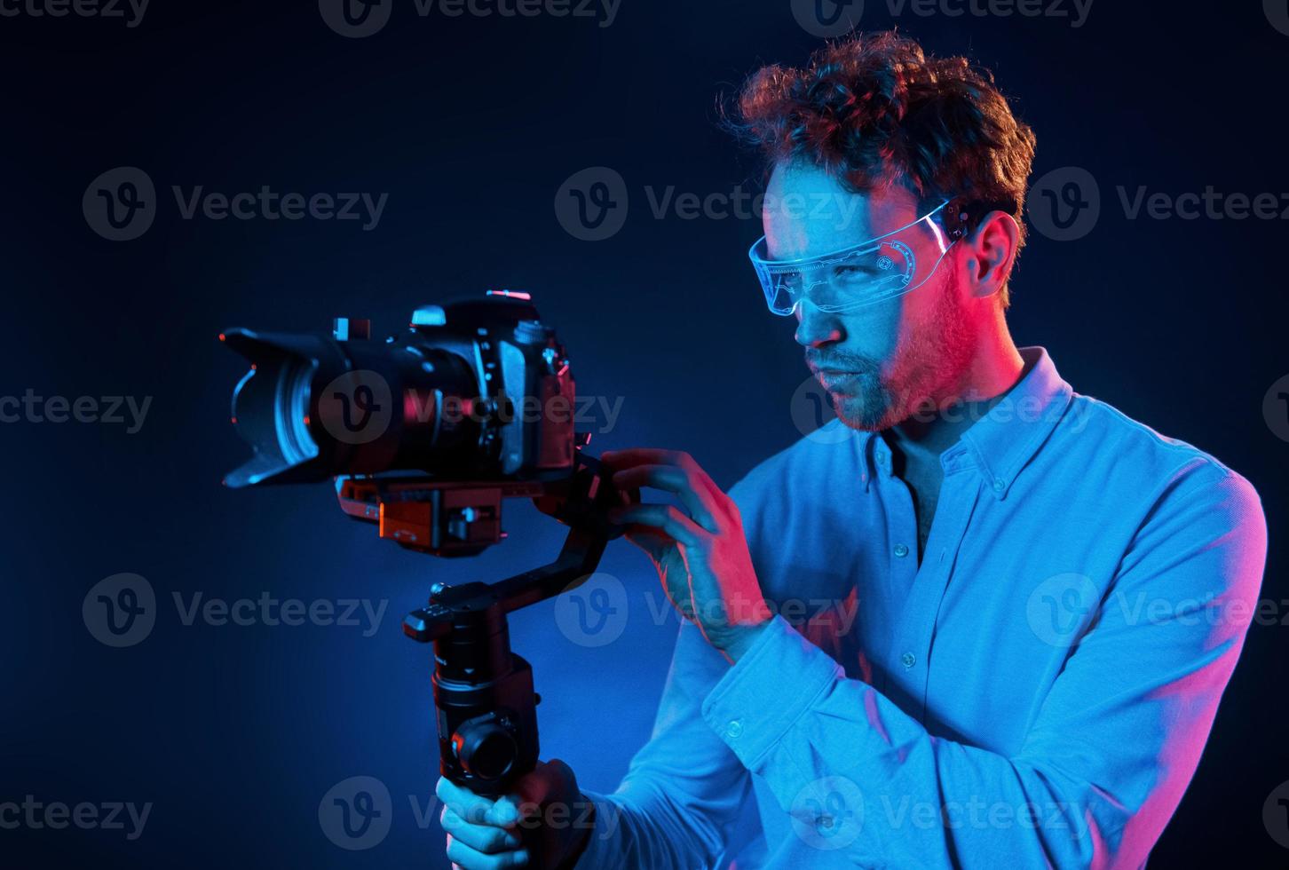 Holds camera. Neon lighting. Young european man is in the dark studio photo