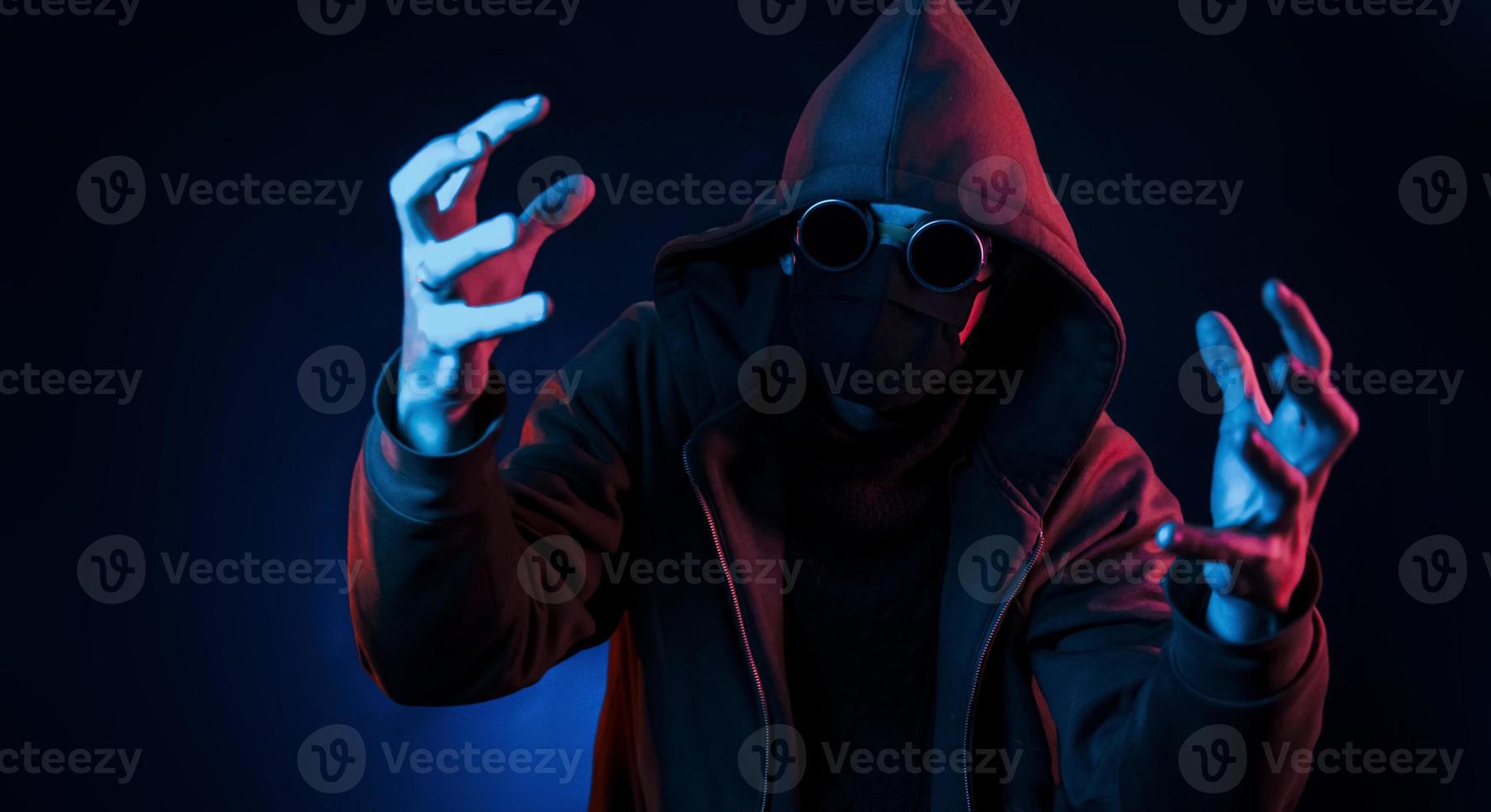 Scary guy in eyewear, mask and hood. Neon lighting. Young european man is in the dark studio photo