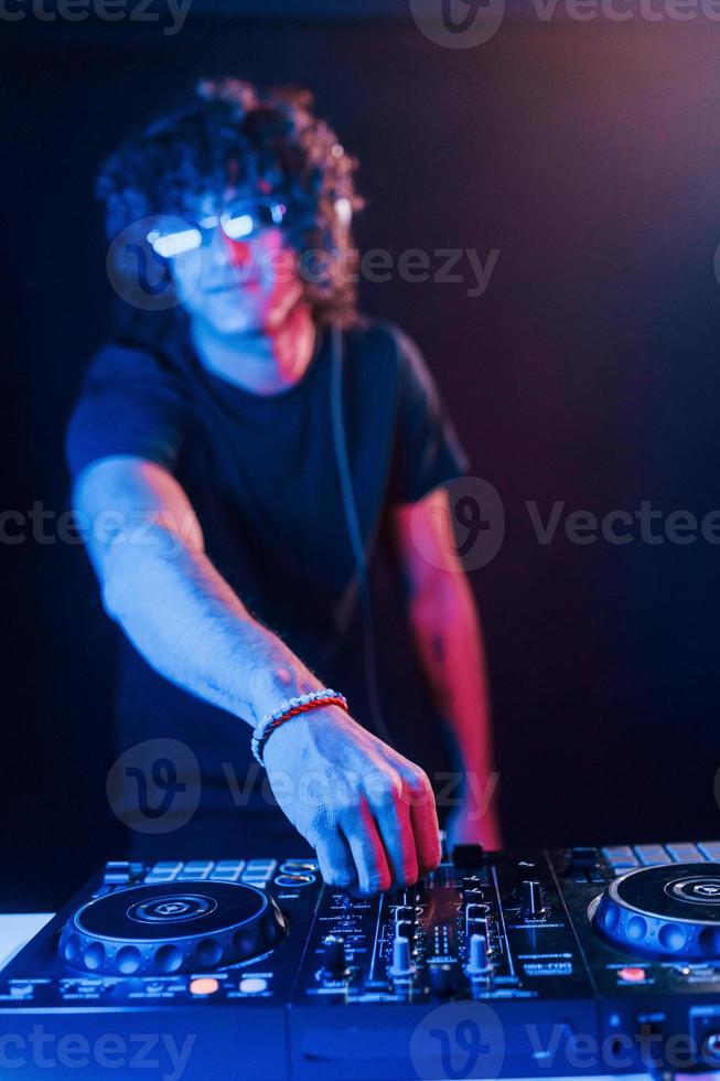 hombre con cabello rizado usando equipo de dj y parado en la oscura habitación iluminada con neón foto