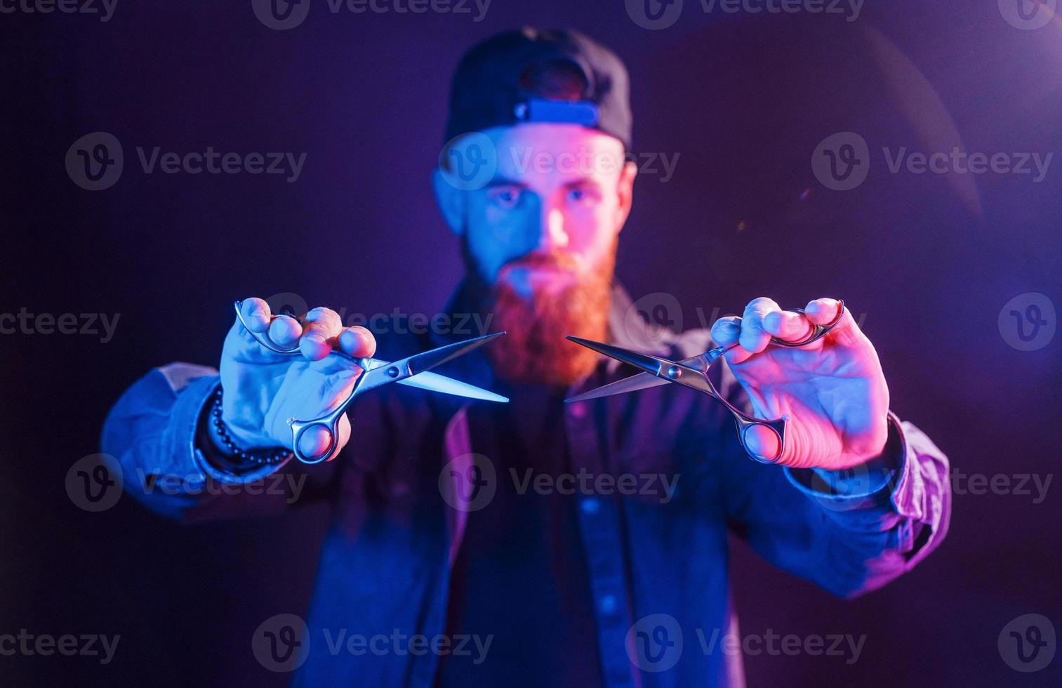 With scissors. Young bearded barber in cap standing in the studio with neon lighting photo