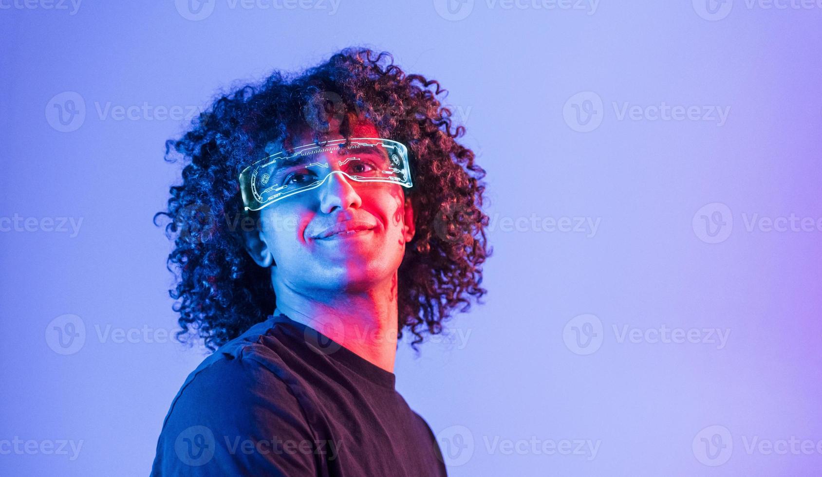 In futuristic glasses. Young beautiful man with curly hair is indoors in the studio with neon lighting photo