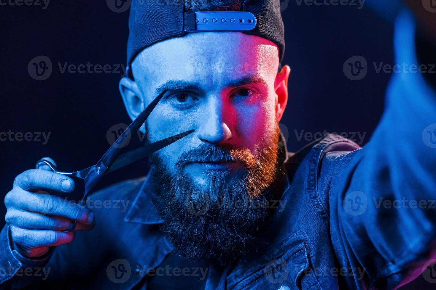 With scissors. Young bearded barber in cap standing in the studio with neon lighting photo