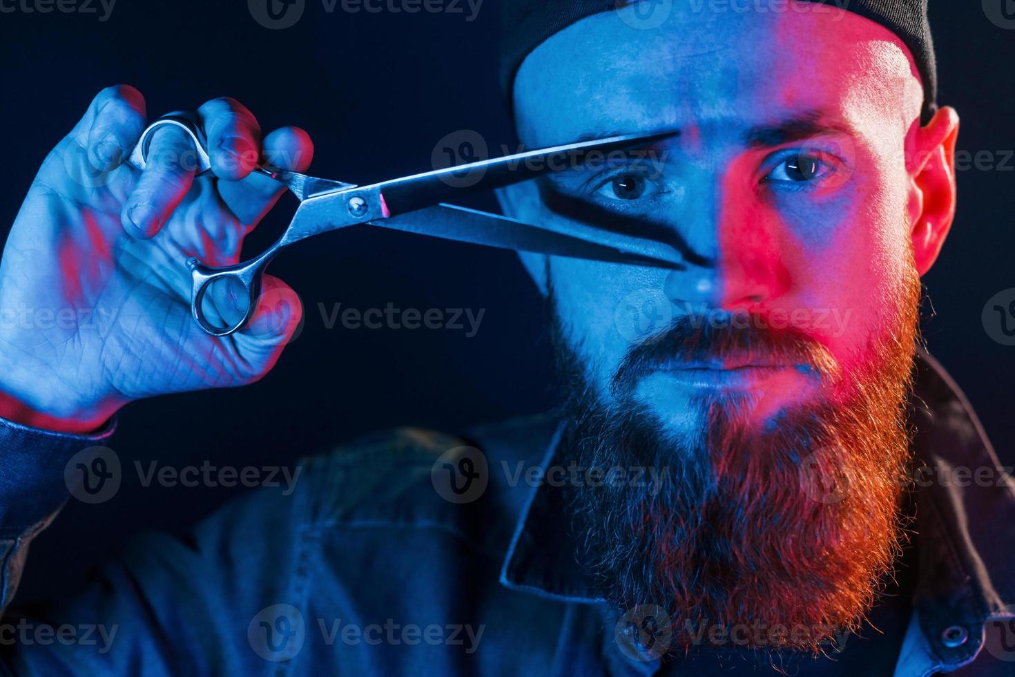 With scissors. Young bearded barber in cap standing in the studio with neon lighting photo