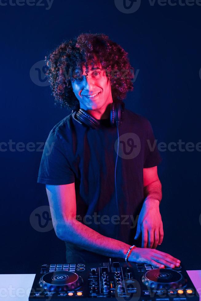 hombre con cabello rizado usando equipo de dj y parado en la oscura habitación iluminada con neón foto