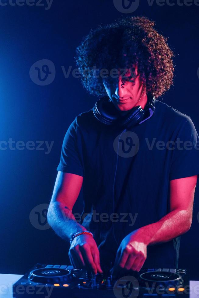 Man with curly hair using DJ equipment and standing in the dark neon lighted room photo