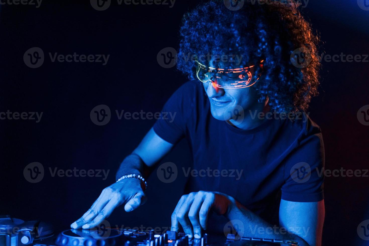 Man with curly hair using DJ equipment and standing in the dark neon lighted room photo