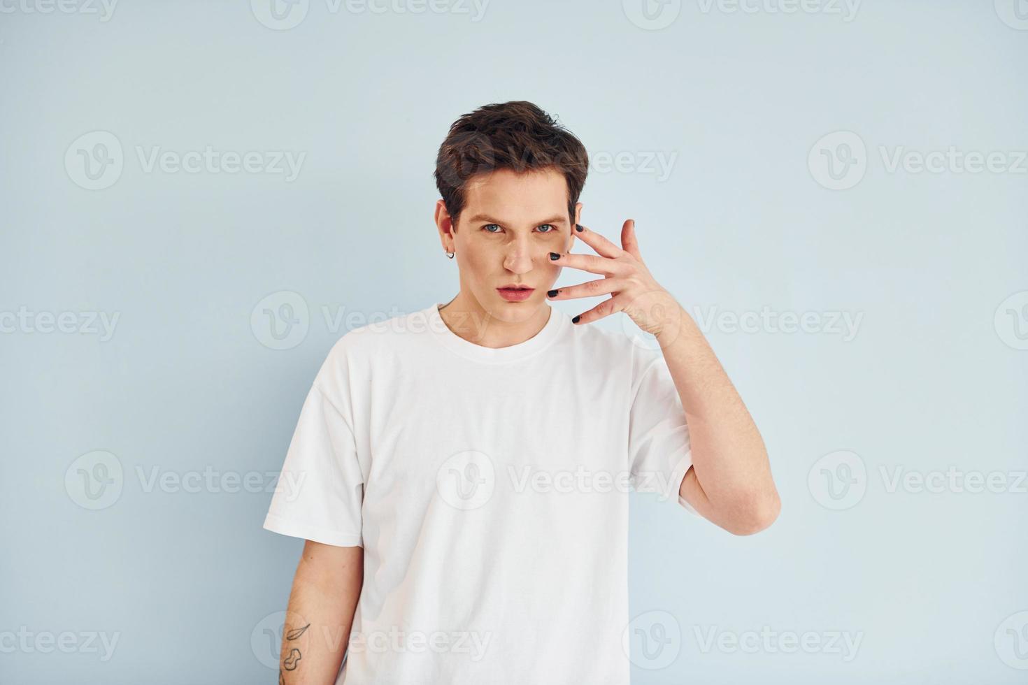 Young gay man is standing in the studio and posing for a camera. In casual white shirt photo