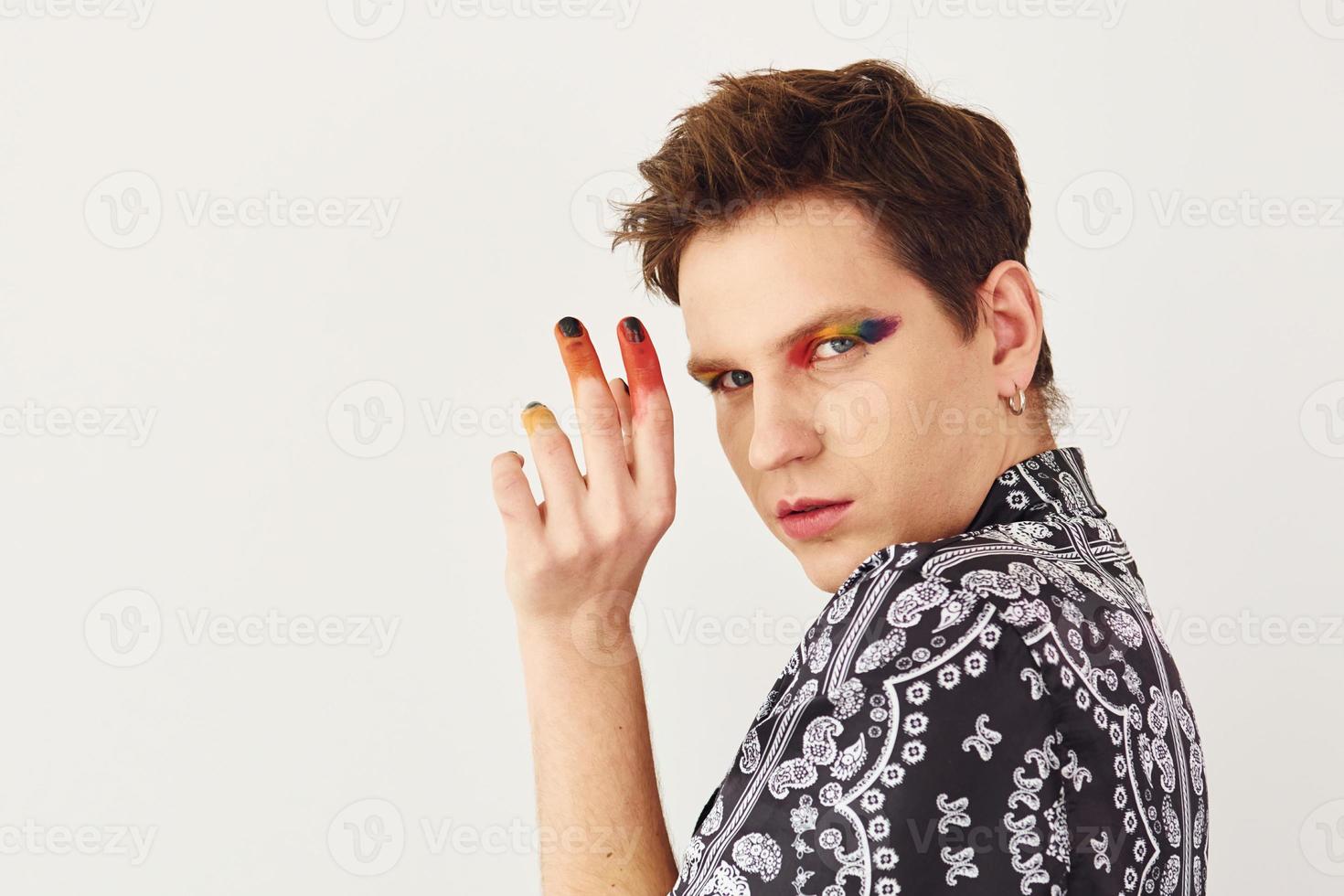 Young gay man is standing in the studio and posing for a camera. Multicolored make up on face fingers photo