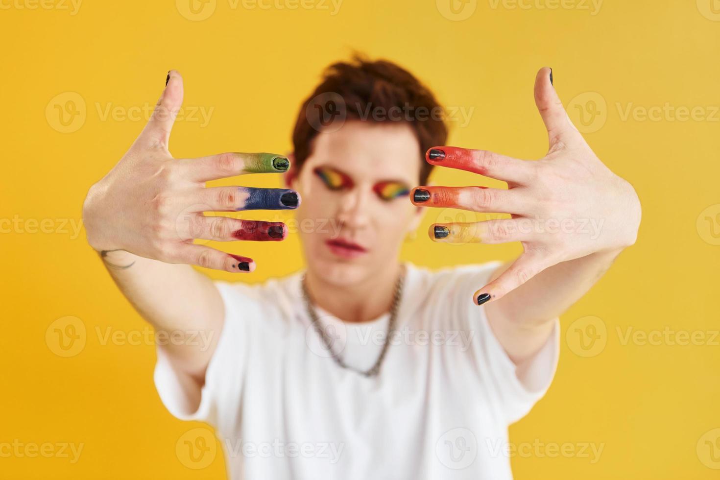 Young gay man is standing in the studio and posing for a camera. Rainbow make up photo