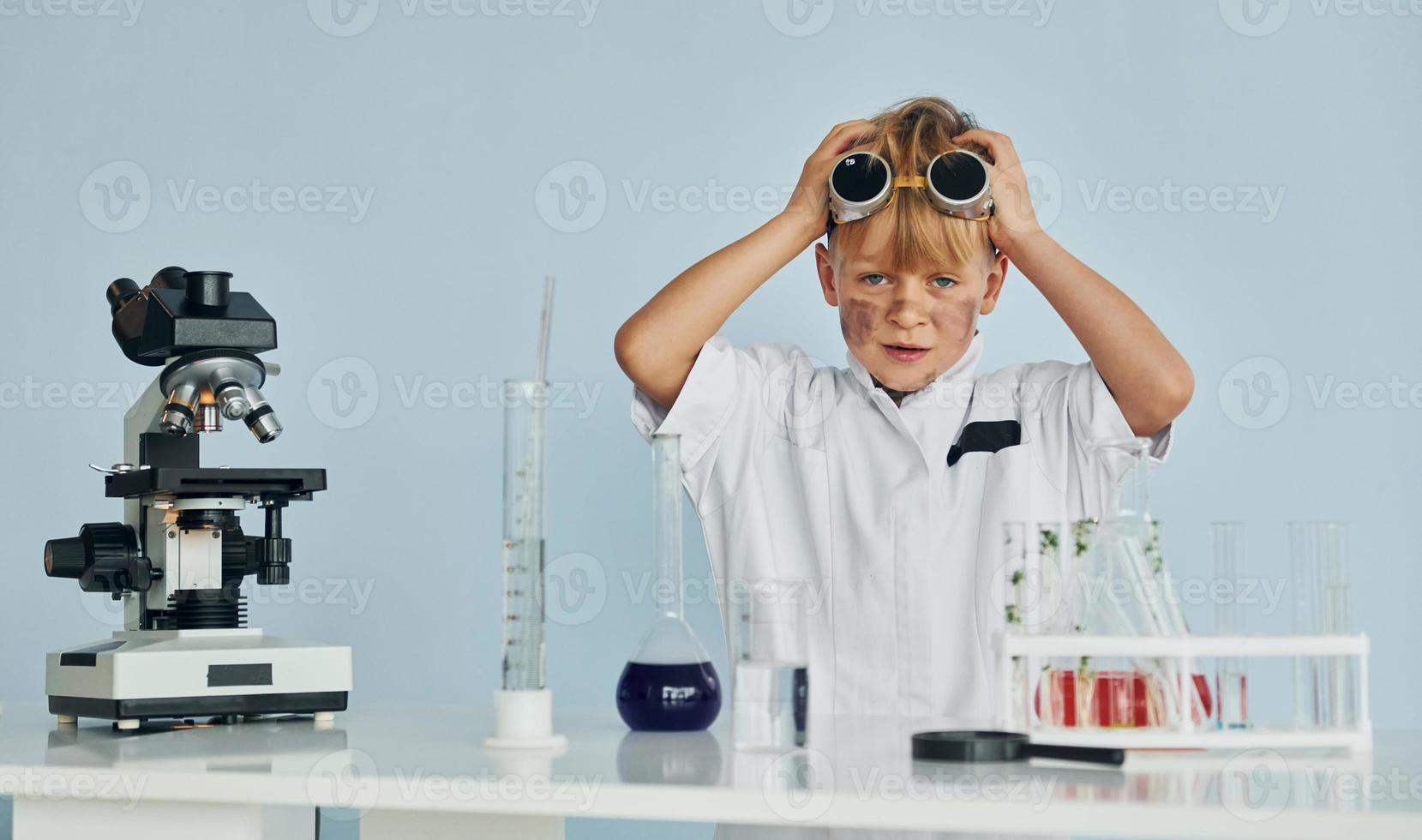 Scared little boy in coat playing a scientist in lab by using equipment photo