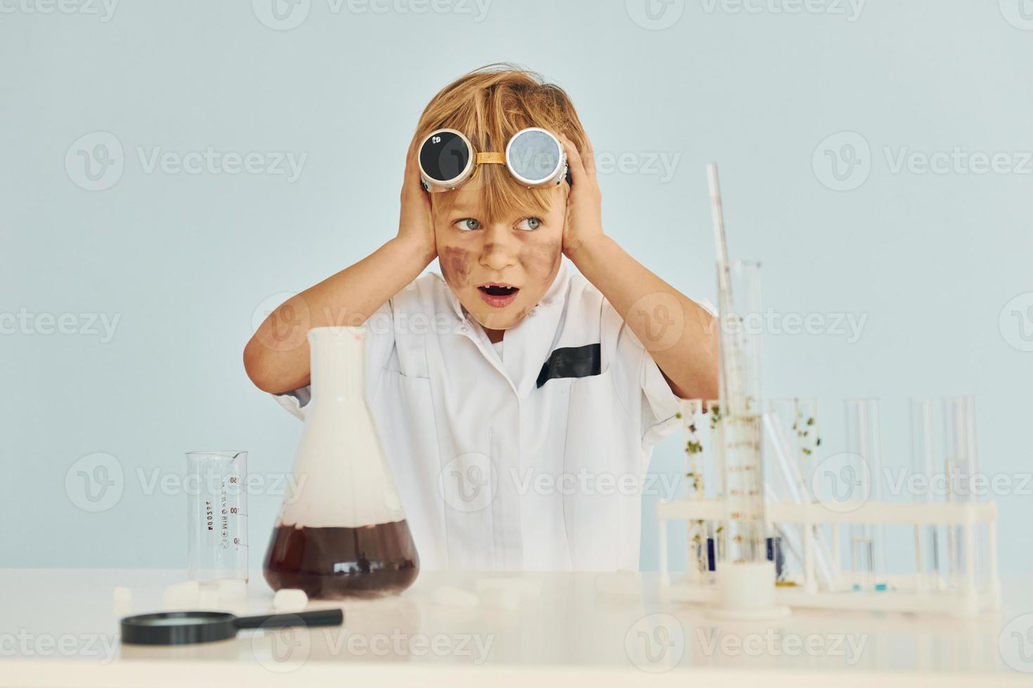 Scared little boy in coat playing a scientist in lab by using equipment photo